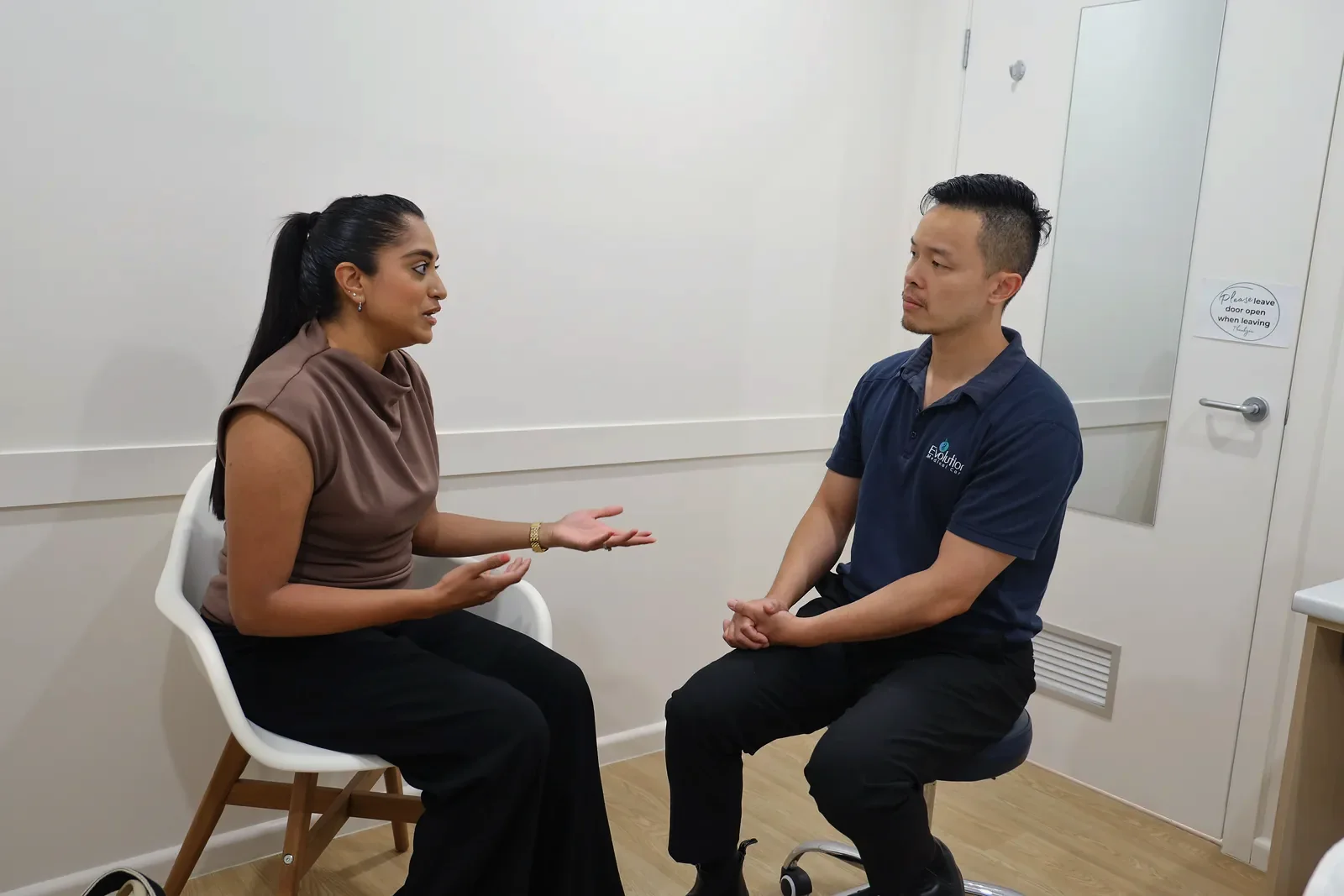 A woman and a practitioner sit in chairs facing each other and talk in a bright consulting room with a closed door behind them.