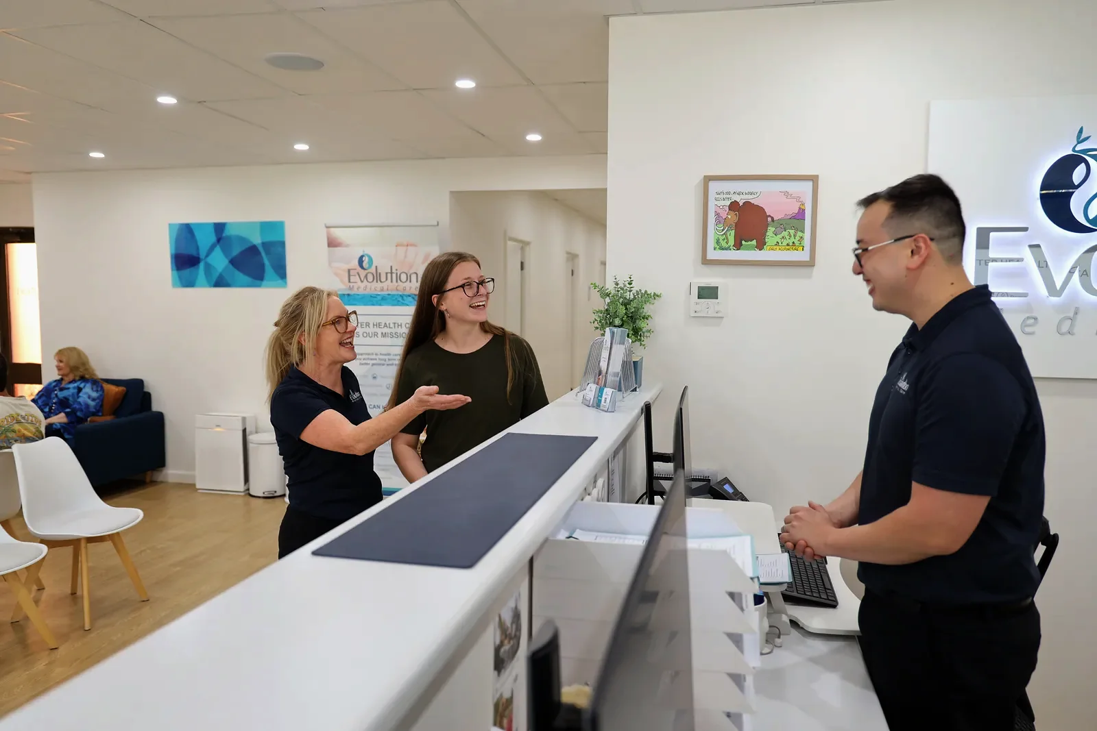 A receptionist, a woman in green and a man in navy at the front desk, with one of them signing a form at the counter.