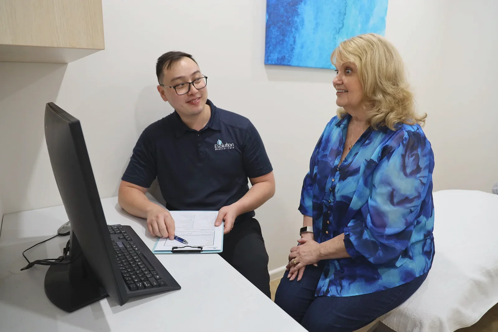 A practitioner in a navy polo shirt holds a clipboard at a desk with a computer while a smiling woman in a blue patterned top sits beside him.