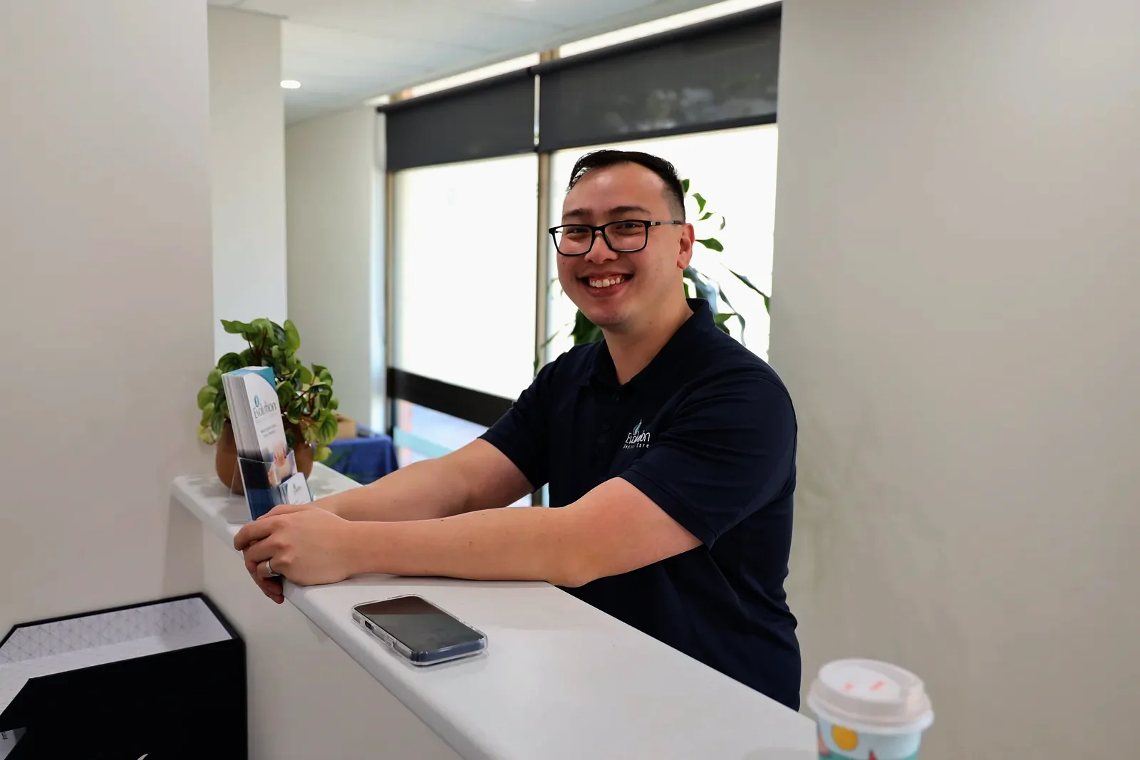A smiling practitioner in a navy polo shirt and glasses leans on the front desk at Evolution Medical Care, with a phone on the counter.