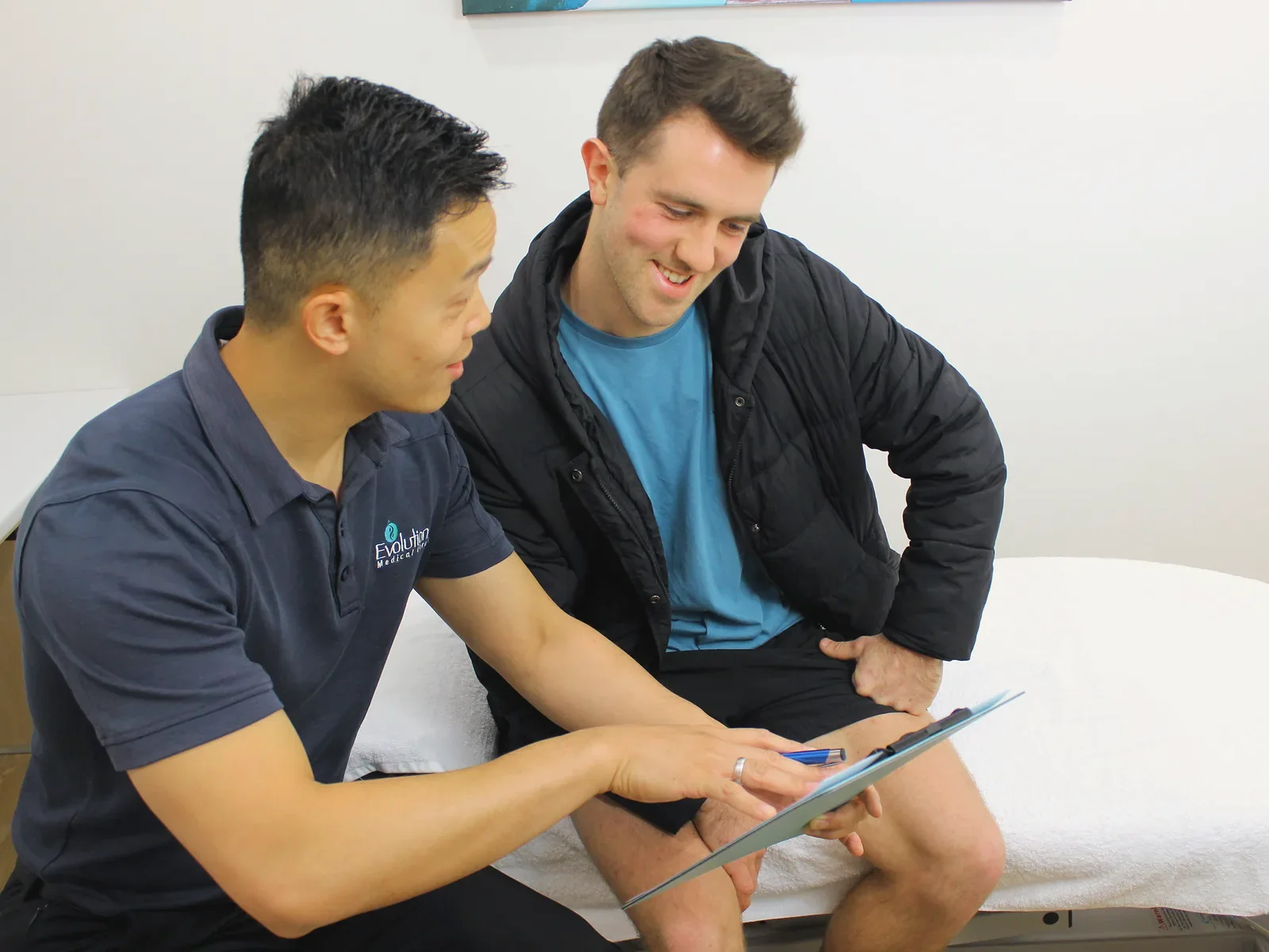 A practitioner in a navy polo shirt points to a clipboard while a smiling man in a dark jacket looks on, seated beside a treatment couch.