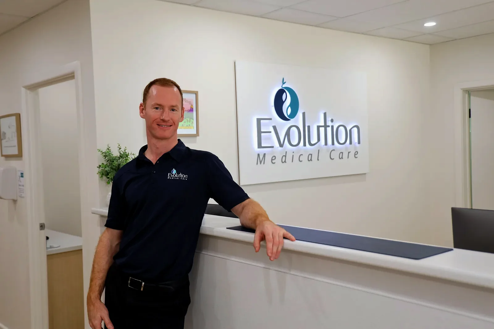 A smiling practitioner in a navy polo shirt leans on the front desk beside the illuminated Evolution Medical Care logo sign.