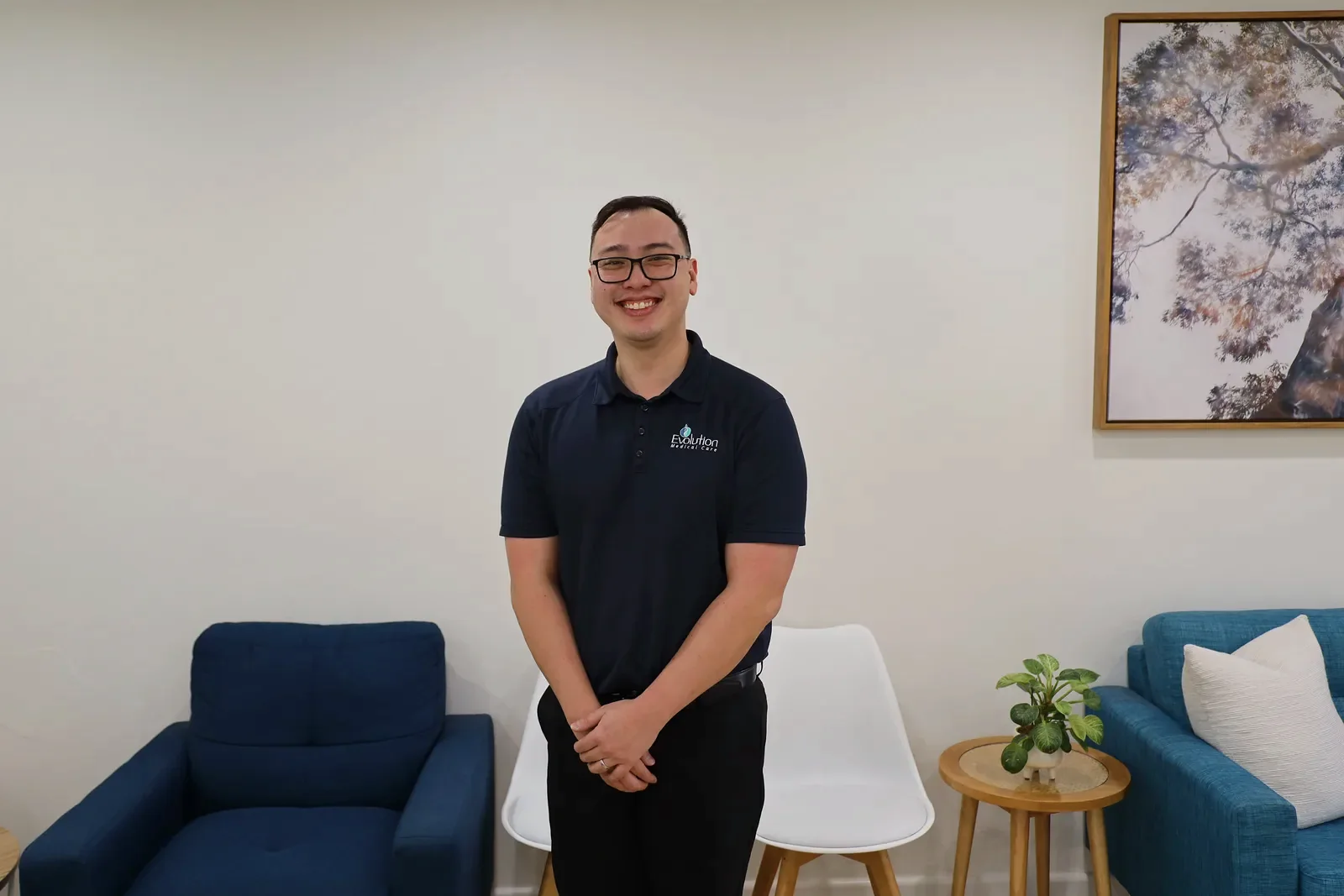 A smiling practitioner in a navy polo stands facing the camera in a waiting room with a navy armchair, white chair and framed blossom print behind him.