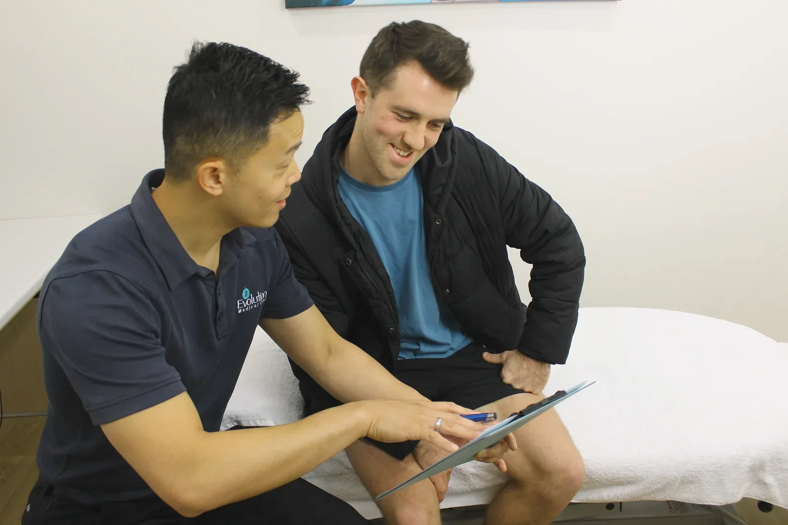 A practitioner points to a tablet while a smiling man in a jacket sits beside him in a consulting room.