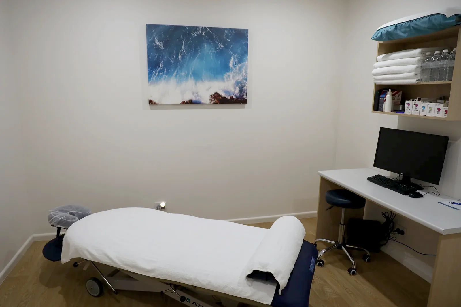 A quiet consulting room with a desk and switched-off monitor, a made-up treatment table, shelved towels and a framed blue artwork.