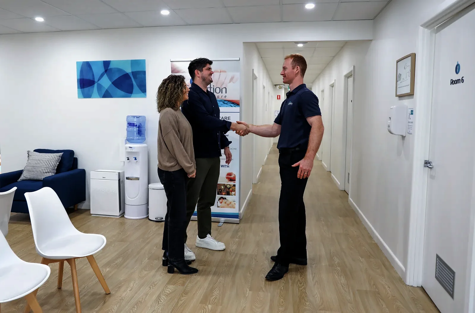 A male staff member shakes hands with a man in a dark jacket in a reception hallway while a woman stands beside them, near a clinic banner.
