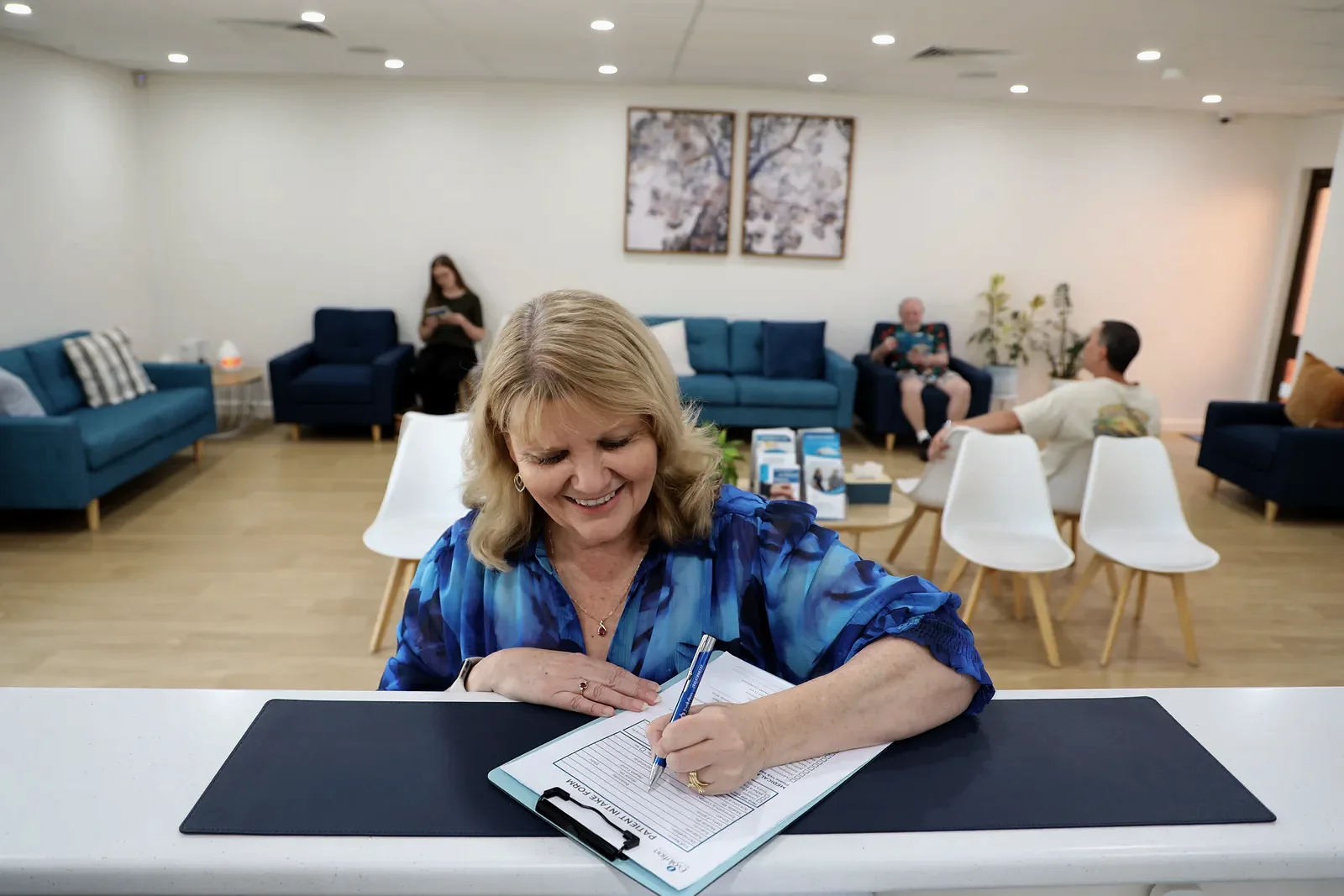 A smiling woman in a blue patterned top writes on a clipboard at the front desk counter, with the waiting room and sofas behind her.