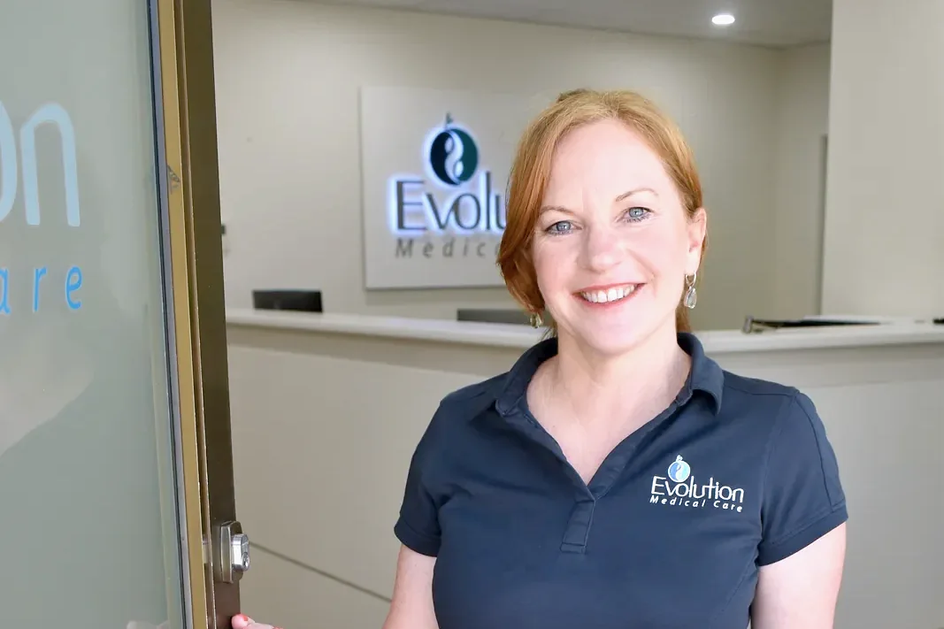 A smiling receptionist in a navy polo holds open a glass clinic door, with the reception desk and logo sign visible behind her.
