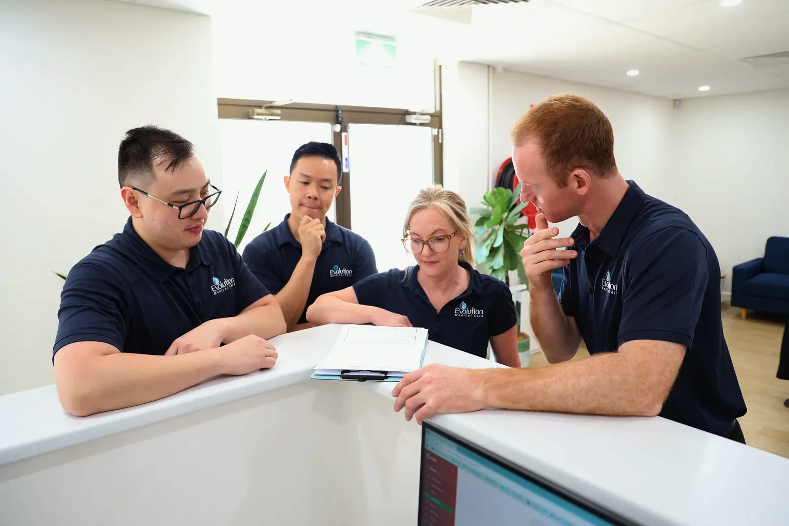 Four Evolution Medical Care staff in navy polo shirts lean over the front desk and look at a clipboard of paperwork together, with a computer screen below.