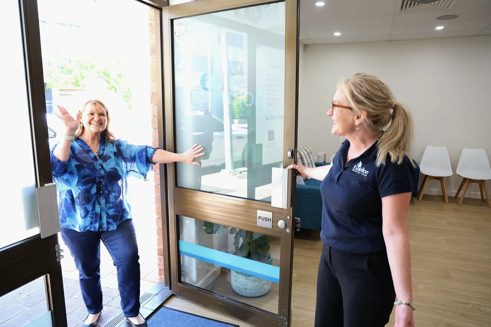 A smiling woman in a blue patterned top waves as she steps through the clinic front door, greeted by a staff member in a navy polo shirt inside.