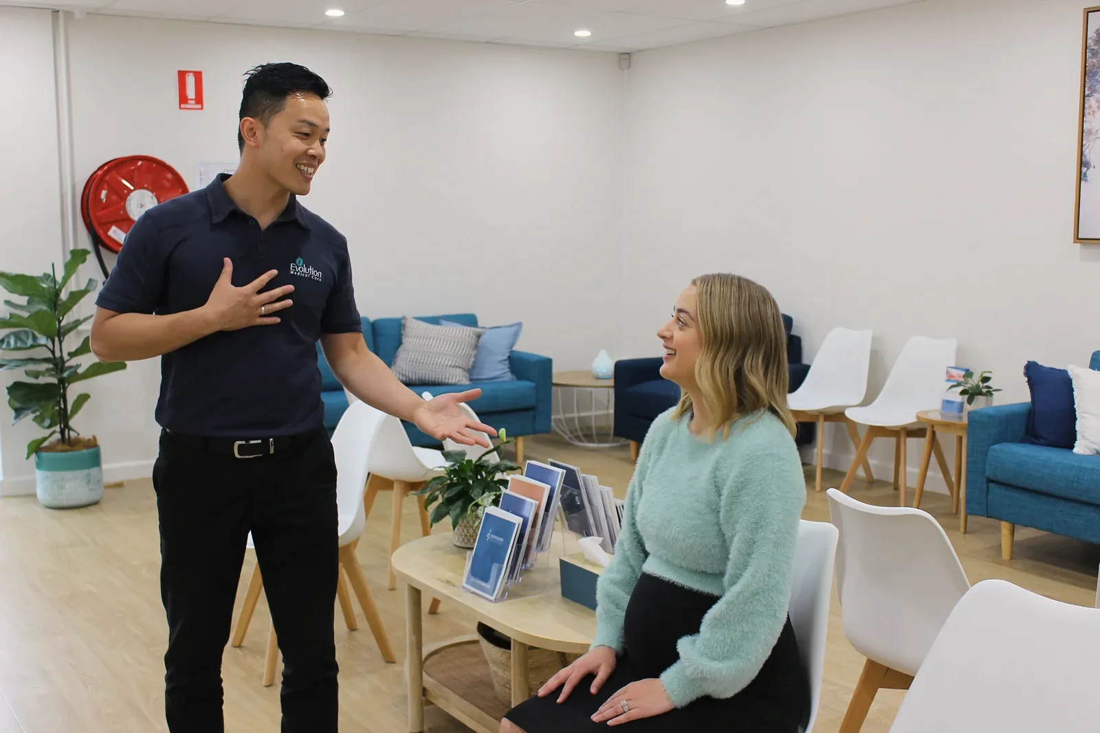 A practitioner places a hand on his chest in a welcoming gesture toward a smiling seated woman in the waiting room.